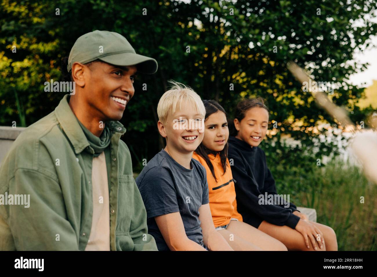 Bambini seduti con un consulente sorridente che indossa un berretto al campo estivo Foto Stock