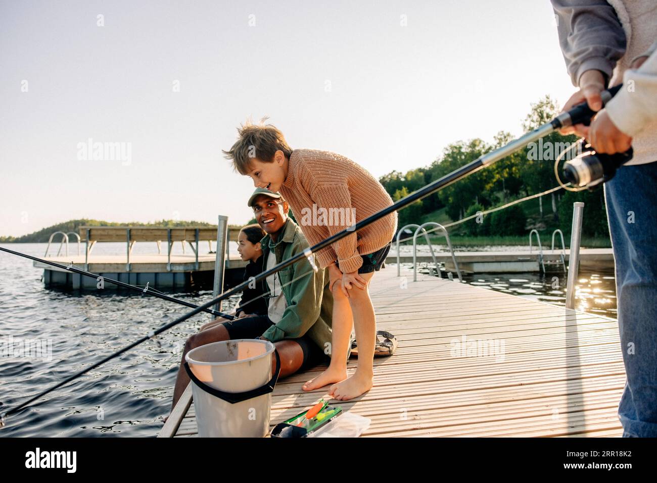 Ragazzo curioso che guarda nel lago con un consulente che pesca sul molo al campo estivo Foto Stock