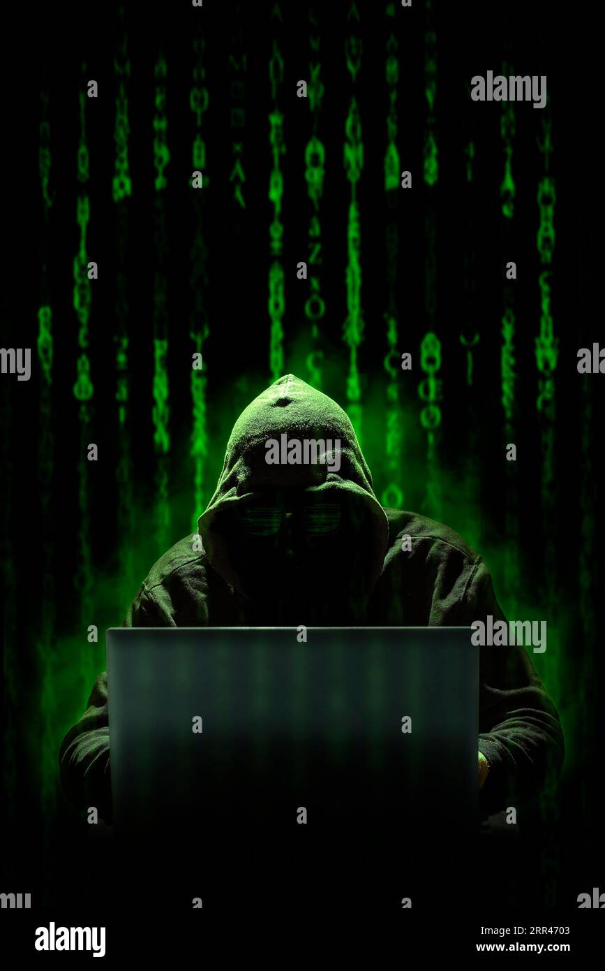 Un hacker incappucciato lavora su un cyber attacco con un notebook, su uno sfondo di codice informatico in stile matrice in cui è possibile vedere un teschio. Crimini informatici. Foto Stock
