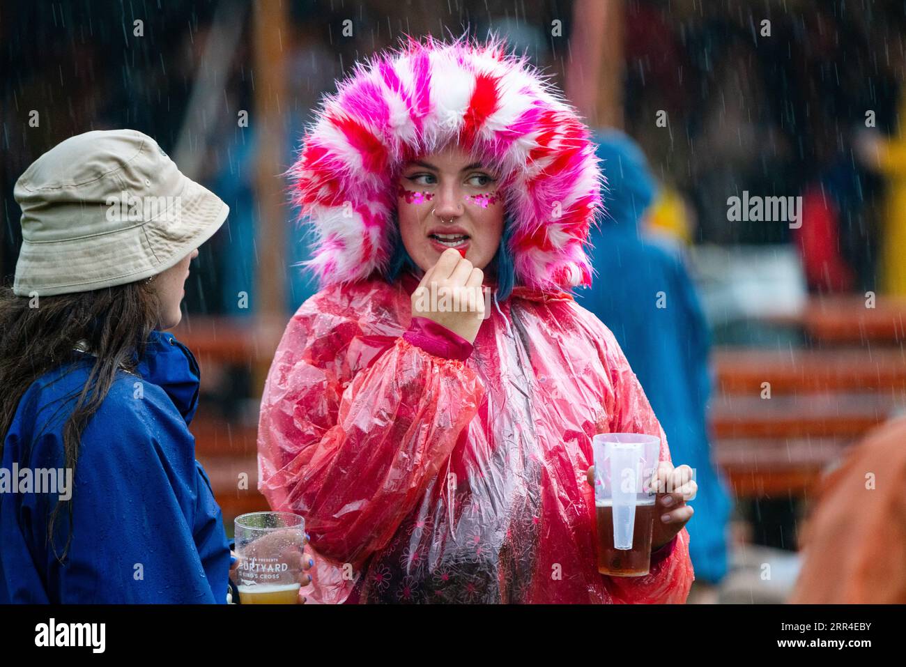 Una donna con cappuccio in pelliccia rosa e poncho antipioggia usa e getta che applica il rossetto al temporale. Green Man Festival, Brecon, Galles, Regno Unito, 2023. Foto Stock