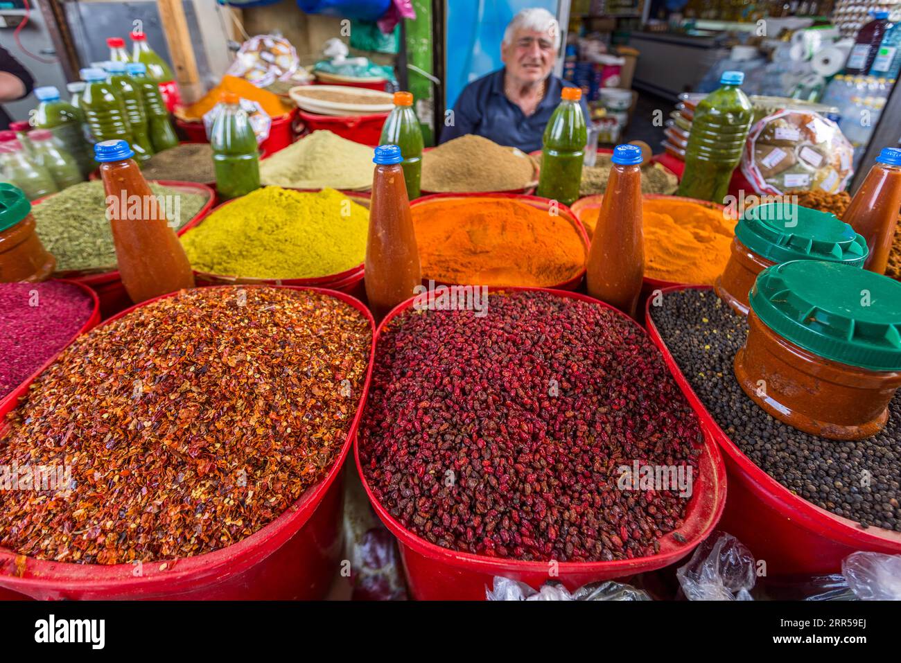 Un commerciante nella sua stalla di spezie e frutta secca al Dezerter Bazaar di Tbilisi, Georgia. In primo piano ci sono grandi ciotole di barbacche essiccate (Berberis vulgaris), utilizzate nella cucina georgiana per condire piatti di carne e salse, nonché altri frutti secchi; dietro il commerciante ci sono polveri di spezie colorate e bottiglie di essenze. Il Dezerter Bazaar è sotto minaccia di demolizione dal 2024. Mikheil Tsinamdzghvrishvili Street, Chughureti, Didube, Tbilisi, 0102, Georgia Foto Stock