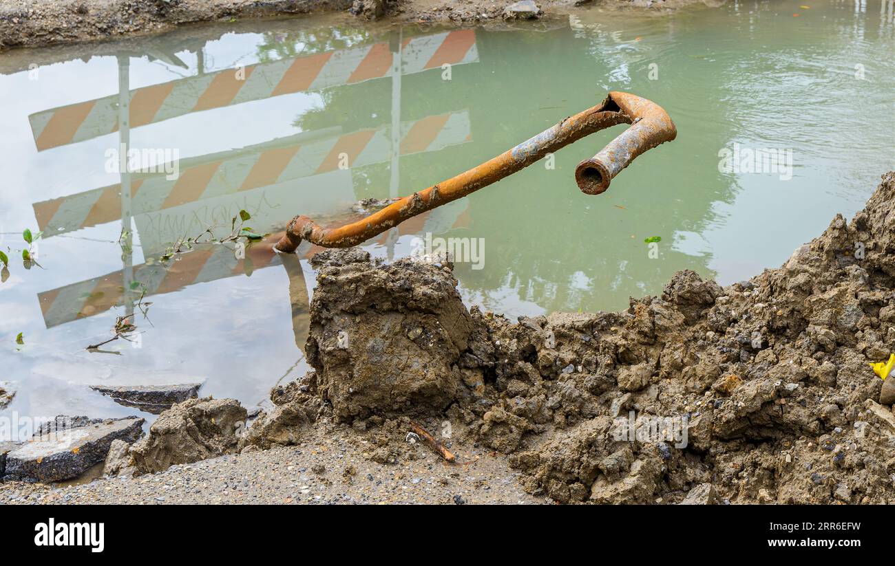 Tubo rotto e arrugginito che si estende da un foro fangoso riempito d'acqua nel sottosuolo di una strada cittadina durante un progetto di riparazione stradale Foto Stock