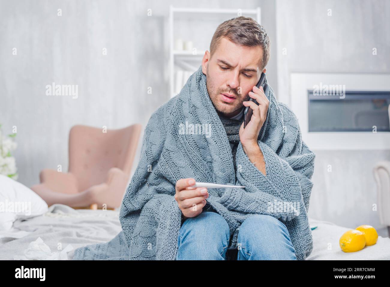 Giovane uomo seduto sul letto avvolto sciarpa che sembra termometro che parla di telefono cellulare Foto Stock