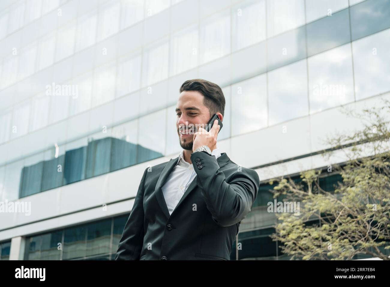 Un giovane uomo d'affari sorridente che si trova in piedi nell'edificio del front Office e parla con un telefono cellulare Foto Stock