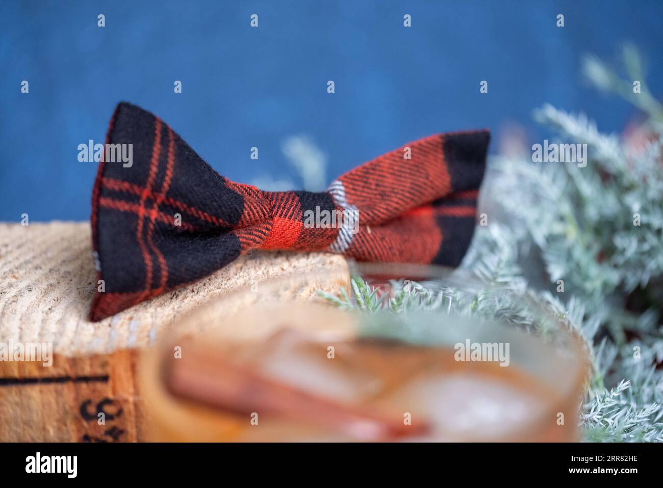 Whisky forte in un bicchiere con ghiaccio e bastoncino di cannella con un ramo di ginepro sullo sfondo, nell'atmosfera natalizia. Fiocco rosso nero a scacchi su un Wo Foto Stock