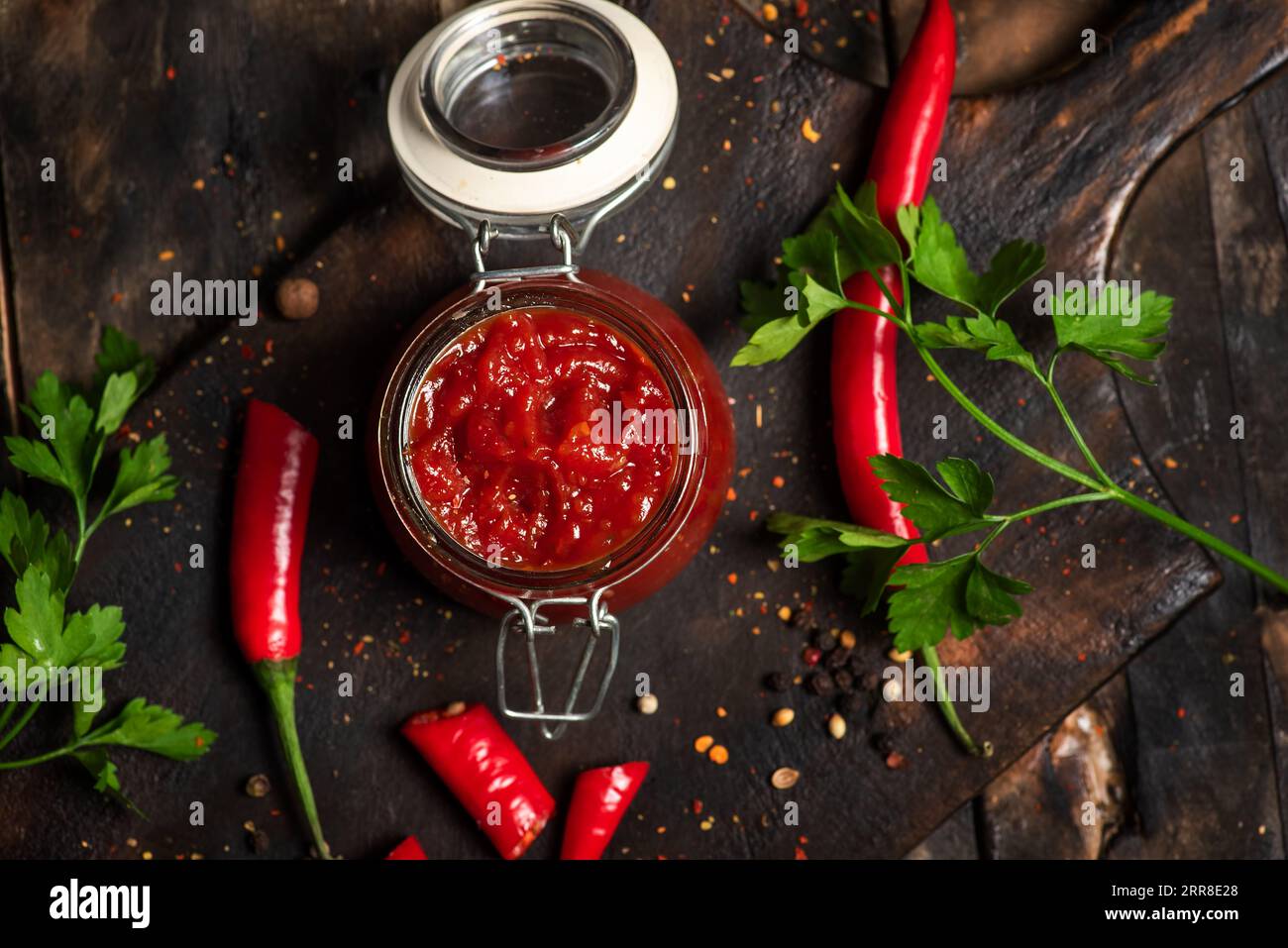 Salsa piccante fatta in casa al peperoncino in un piccolo vaso di vetro su un tavolo da cucina in legno scuro, con peperoni rossi caldi e prezzemolo Foto Stock