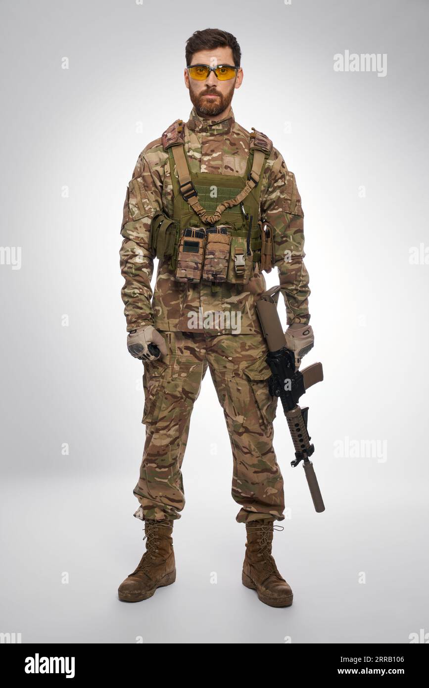 Seri soldato maschio in armatura che tiene il fucile, mentre posa in interni. Vista frontale del militare dai capelli scuri mimetizzato con l'arma, fotocamera dall'aspetto, con sfondo grigio. Concetto di forza militare. Foto Stock