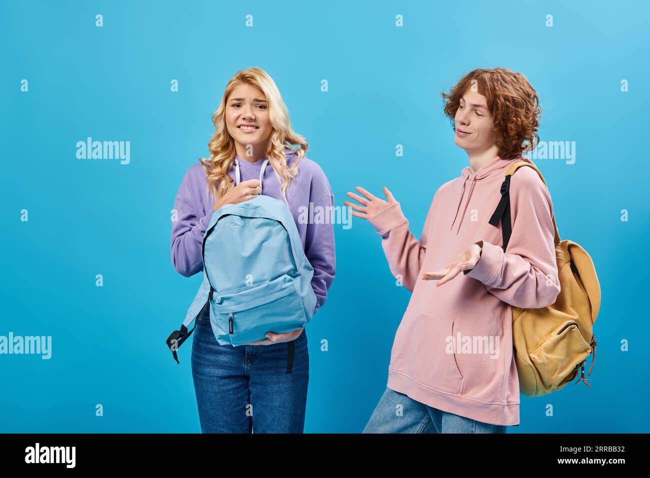 allegro adolescente a testa rossa che si muove vicino a una ragazza scontenta che tiene uno zaino pesante sul blu Foto Stock