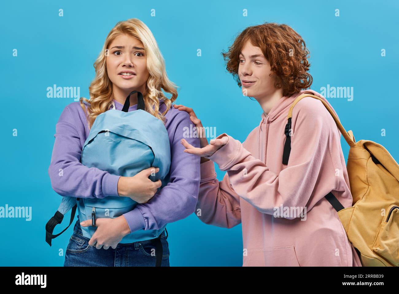 sorridente ragazzo adolescente a testa rossa che si muove vicino alla fidanzata scioccata che tiene uno zaino pesante sul blu Foto Stock