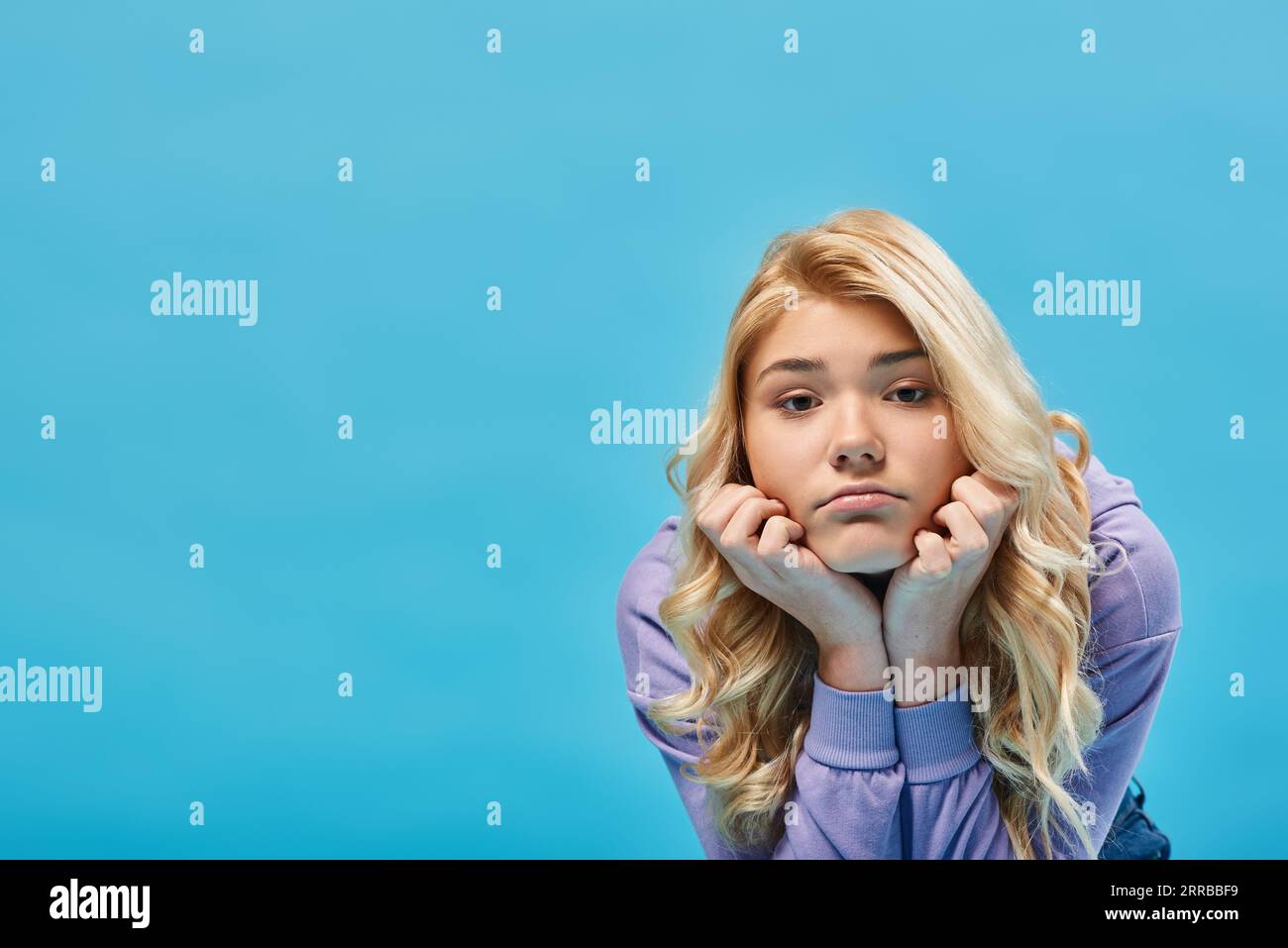 ragazza adolescente bionda frustrata con felpa con cappuccio che tiene le mani vicino al volto e guarda la fotocamera su blu Foto Stock