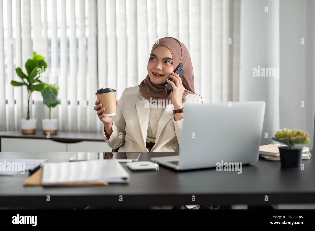 Una donna d'affari musulmana asiatica felice e professionale o un'imprenditrice che indossa un hijab parla al telefono mentre le beve un caffè mattutino Foto Stock