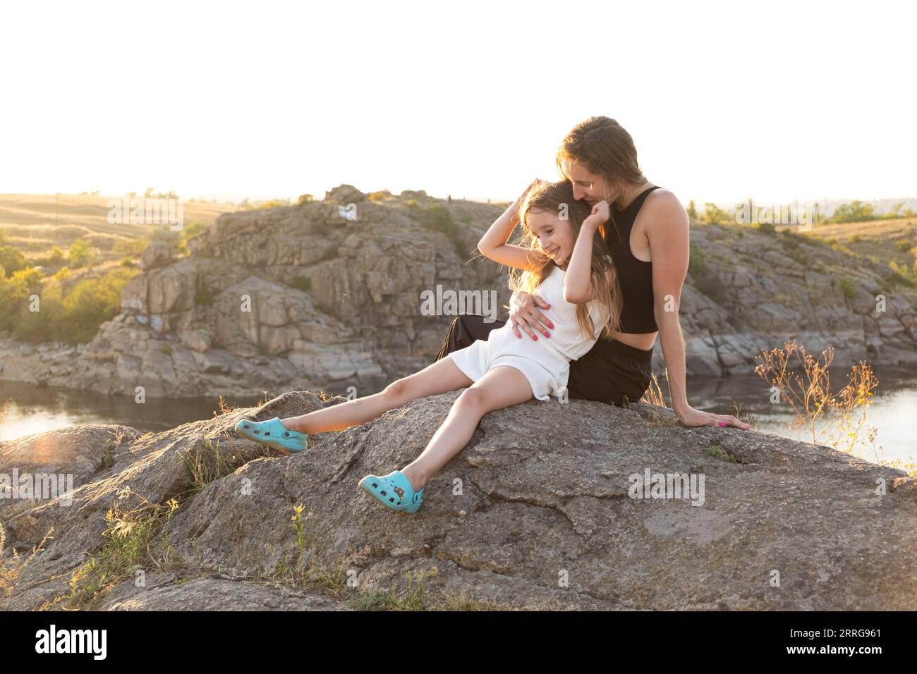 Mamma e figlia felici che si rilassano sulle rocce nella natura sullo sfondo del fiume. Buona maternità, festa della mamma Foto Stock