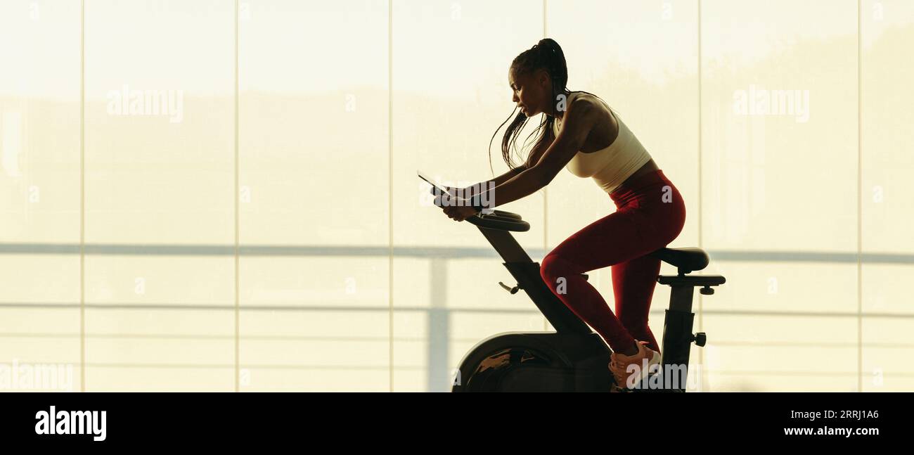 La donna nera rimane in pista con il suo allenamento in palestra. Atleta che partecipa a una lezione di ciclismo online e che usa la sua cyclette per seguirla Foto Stock