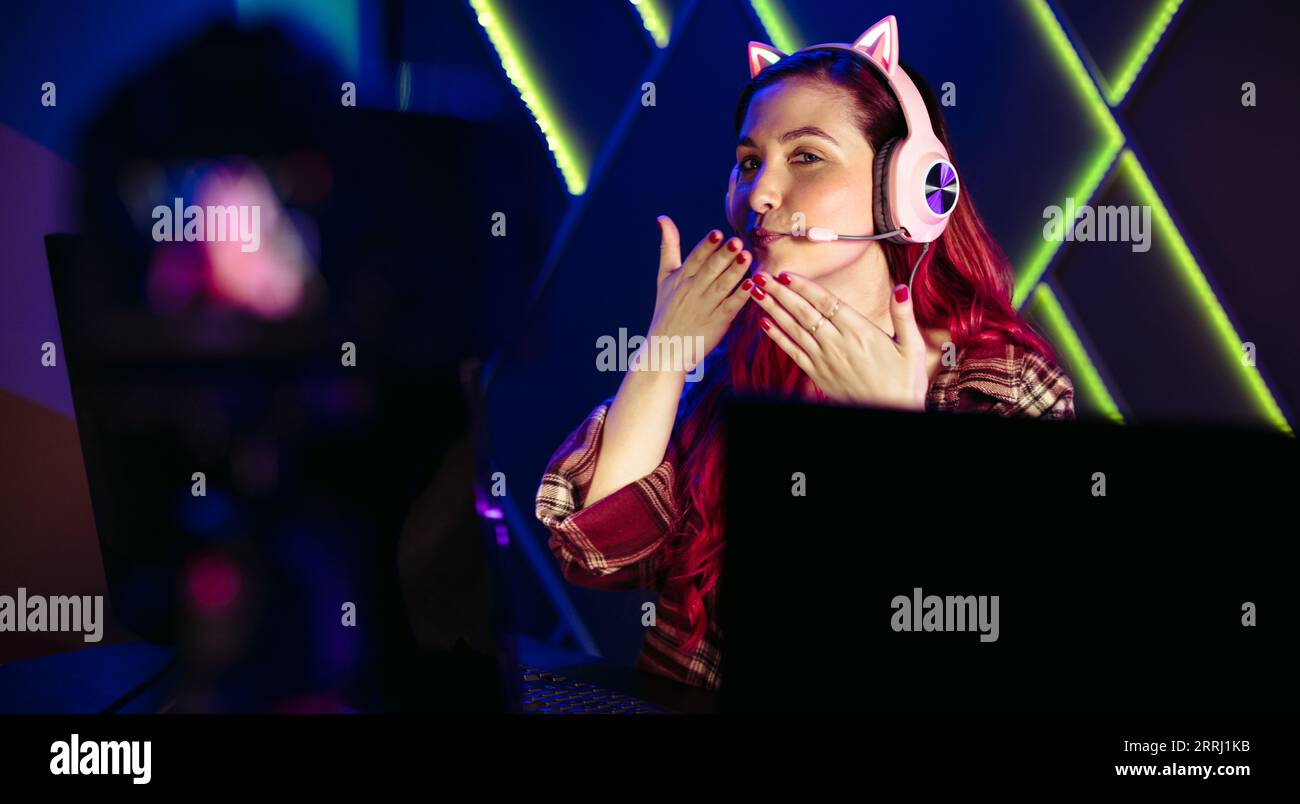 Un'influencer di gioco femminile lancia un bacio mentre registra un live streaming del suo gameplay con una telecamera. Donna che crea contenuti di gioco in streaming online Foto Stock