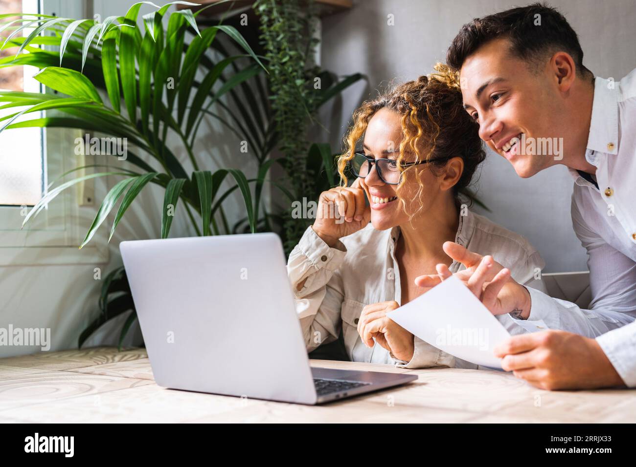 Due persone che lavorano insieme con il computer alla scrivania nel luogo di lavoro dell'ufficio domestico. Un giovane uomo e una donna adulta sorridono e amano le piccole imprese. Imprenditore con assistente che utilizza il computer wireless online Foto Stock