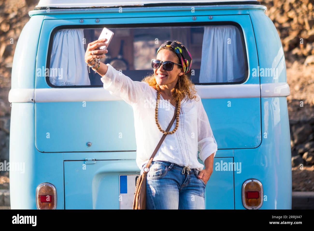 Una giovane donna adulta che scatta selfie con il telefono contro un furgone classico blu. Concetto di persone di viaggio e stile di vita hippy. Una donna viaggiatrice che si appresta a visitare la destinazione. Indossa abiti casual alla moda Foto Stock