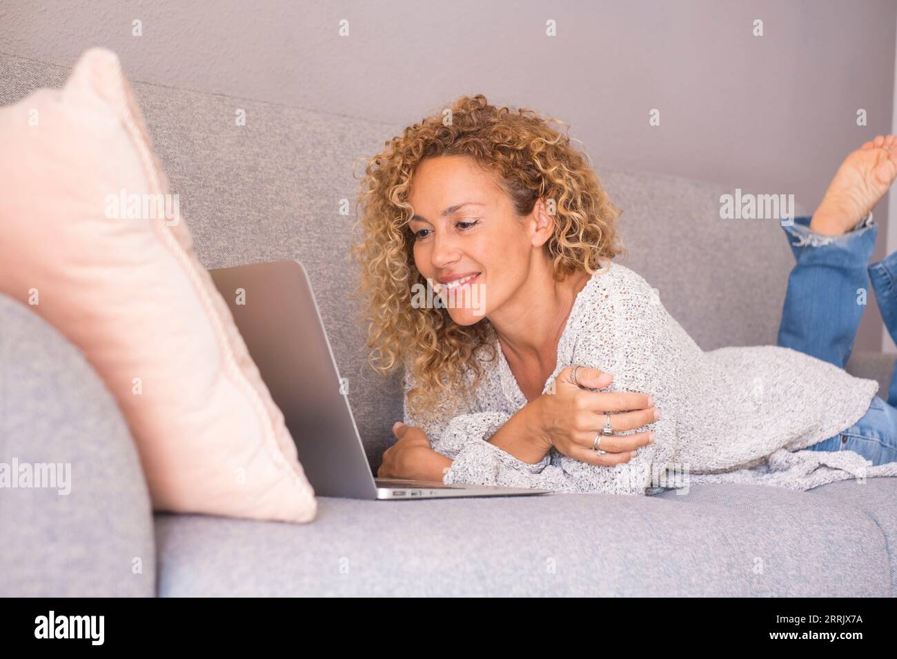Ritratto di una giovane donna adulta felice usando il computer sdraiato sul divano a casa in un ambiente interno rilassato e divertente. Persone che lavorano da freelance comodamente sul divano. Navigare in rete sui social network Foto Stock