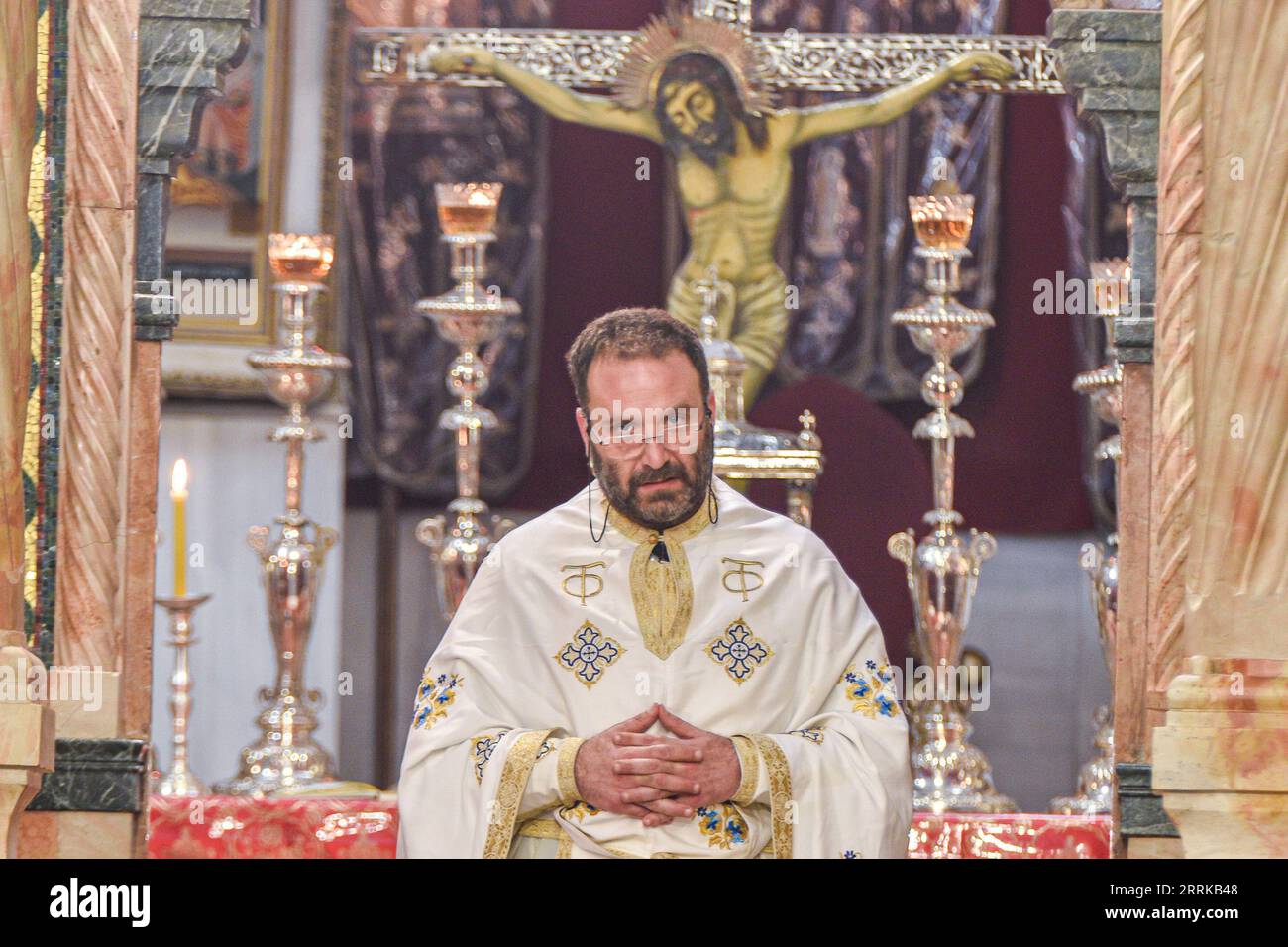 Un uomo di mezza età che indossa occhiali è seduto su un altare in un ambiente religioso, con crocifissi sullo sfondo Foto Stock