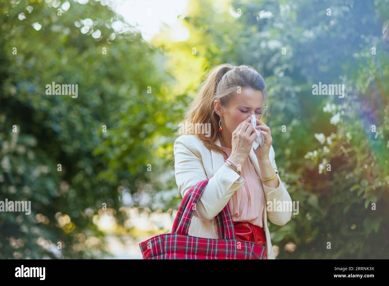 giovane donna in abito rosa e giacca bianca in città con tovagliolo e borsa rossa ha un attacco allergico. Foto Stock