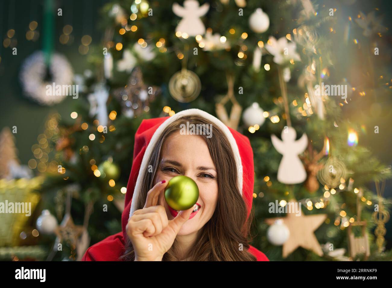 Natale. Donna elegante sorridente con cappello di Babbo Natale e palla di Natale vicino all'albero di Natale nella casa moderna. Foto Stock