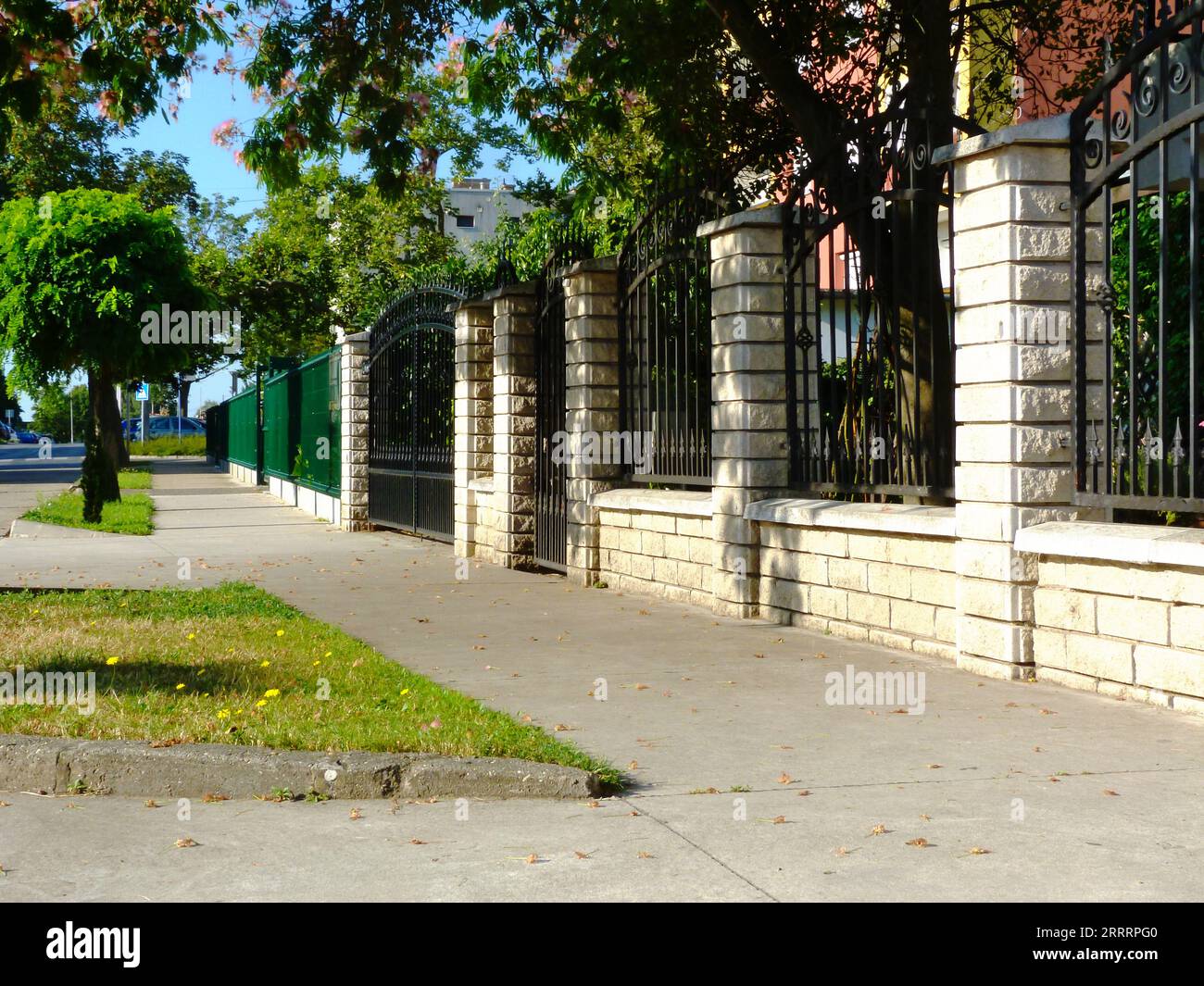 recinzione in ferro battuto nero tra i pilastri in pietra bianca. lussureggiante giardino verde. concetto di proprietà domestica. marciapiede in calcestruzzo. erba verde. alberi lungo la strada Foto Stock