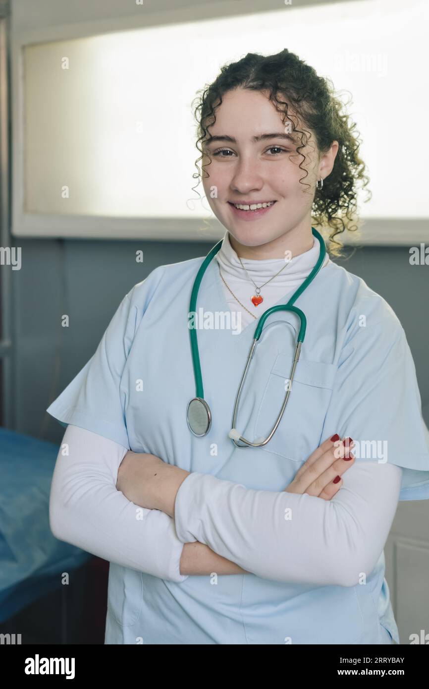 Sorridente giovane medico infermiera medica che indossa cappotto bianco e stetoscopio intorno al collo in piedi in clinica privata con le braccia incrociate guardando camer Foto Stock