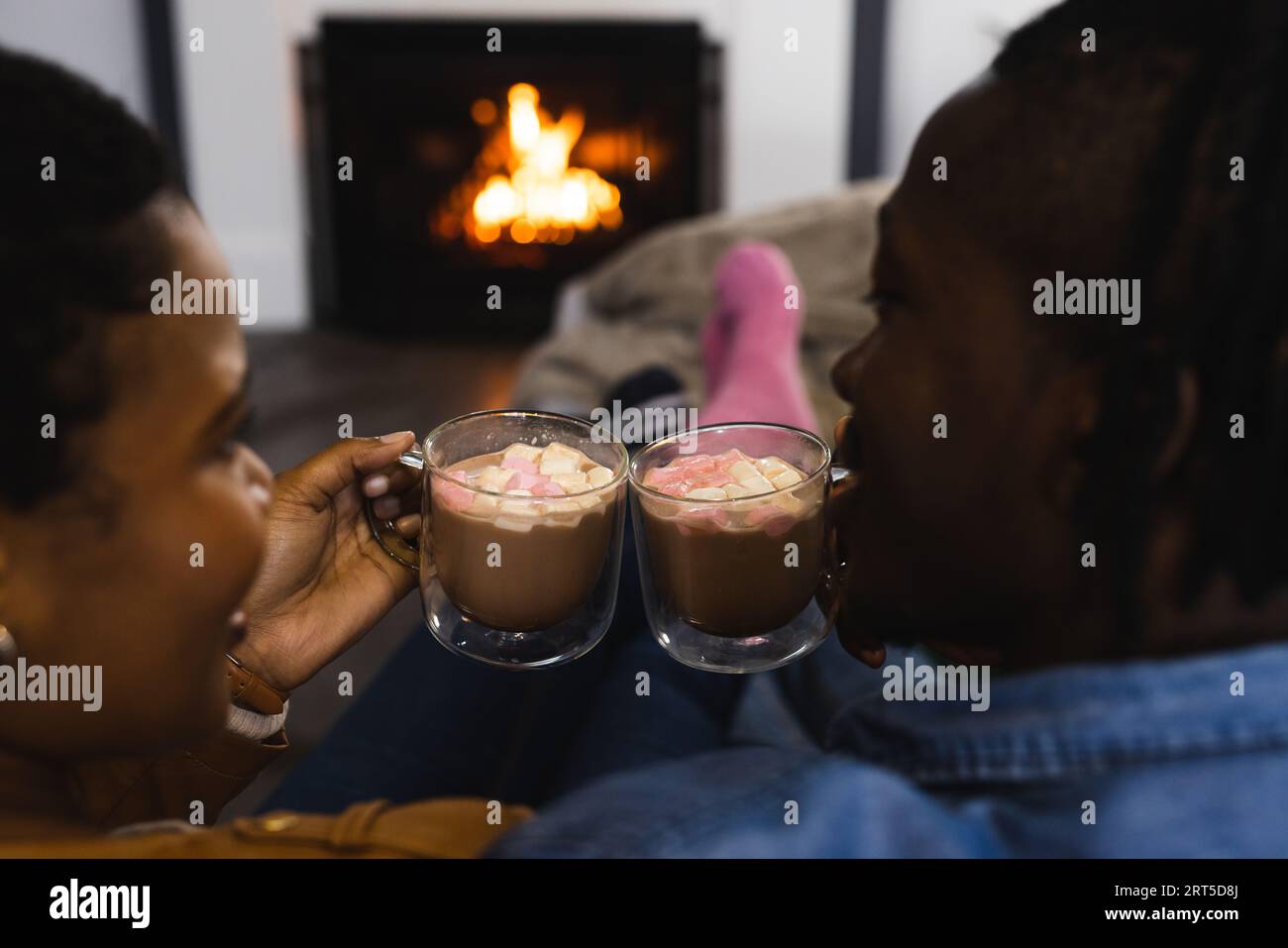 Felice coppia afro-americana che si rilassa a casa davanti al fuoco, facendo un brindisi con cioccolata calda Foto Stock