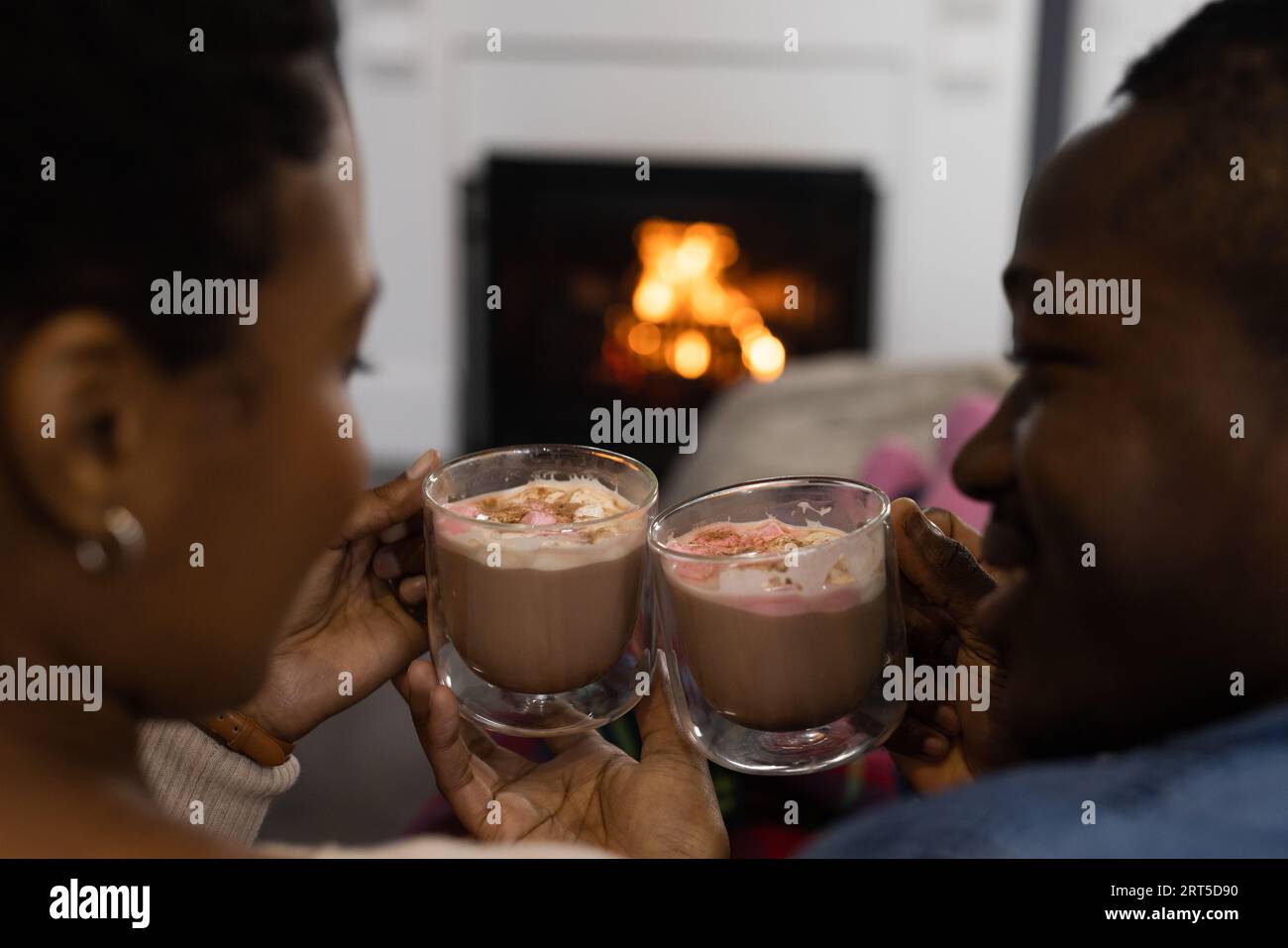 Felice coppia afro-americana che si rilassa a casa davanti al fuoco, facendo un brindisi con cioccolata calda Foto Stock