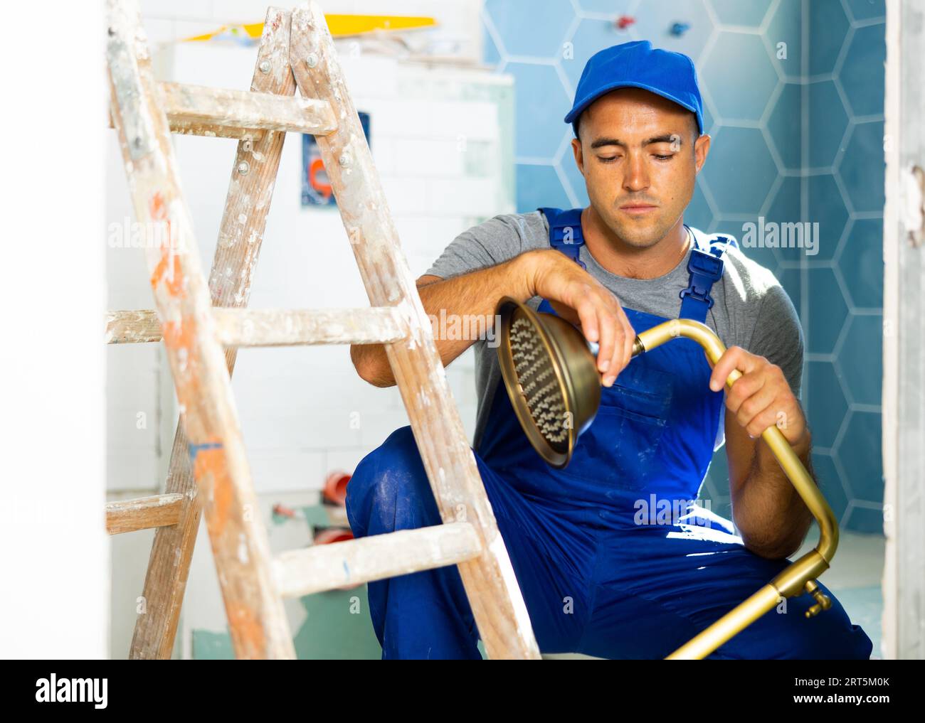 Operaio esperto che indossa tute blu da cantiere seduta e soffione doccia in bagno Foto Stock