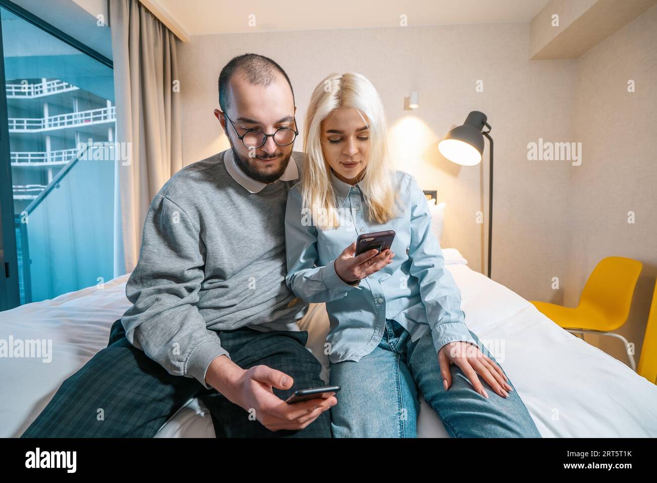 Giovani coppie che guardano il loro telefono mentre sono sedute a letto a casa, trascorrendo del tempo insieme a moglie e marito. Foto Stock