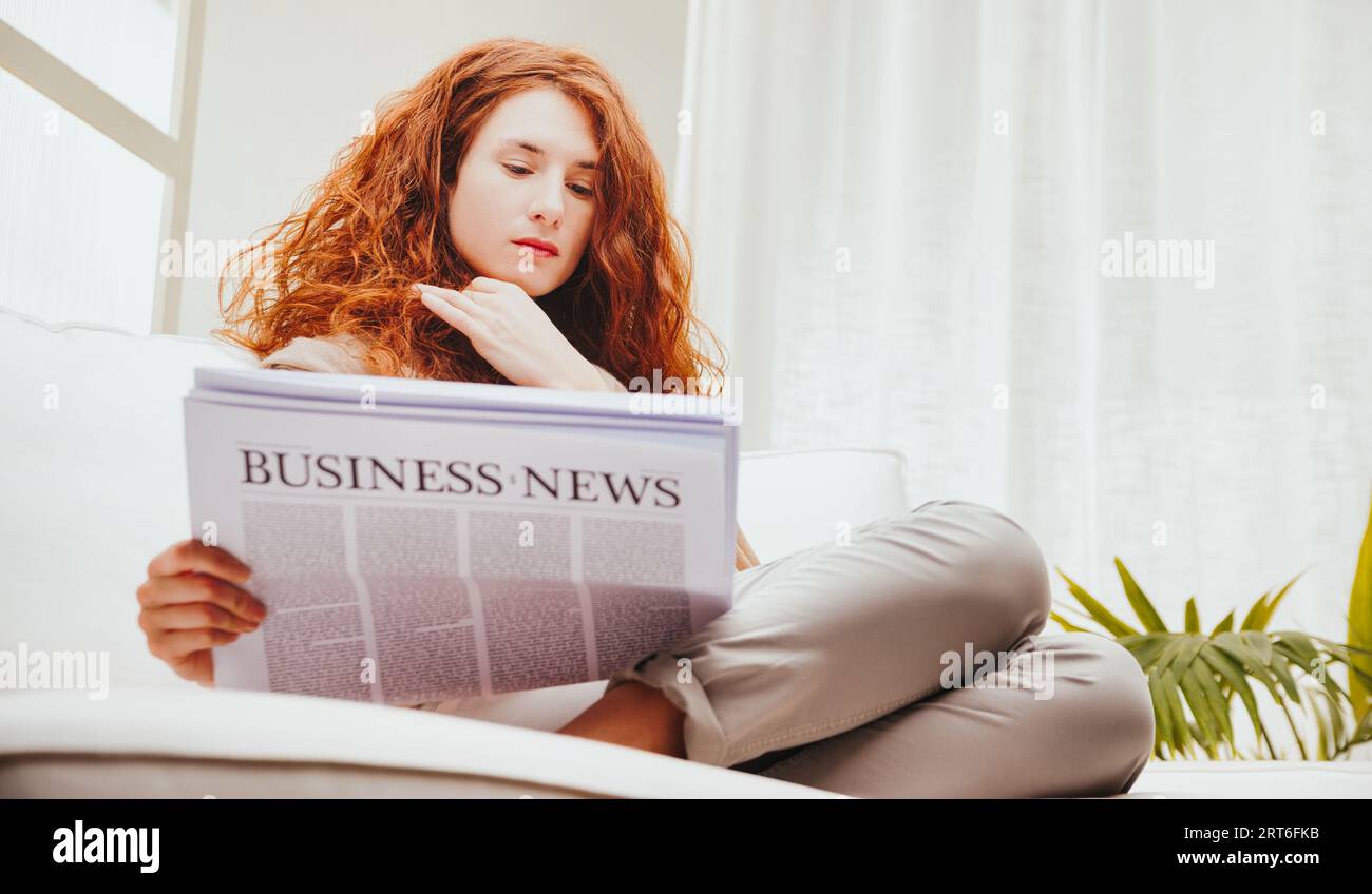 Unendo la raffinatezza al relax, una donna studia i titoli aziendali. Con le gambe che ricordano una sirena, la brillante testa rossa ti piacerà Foto Stock