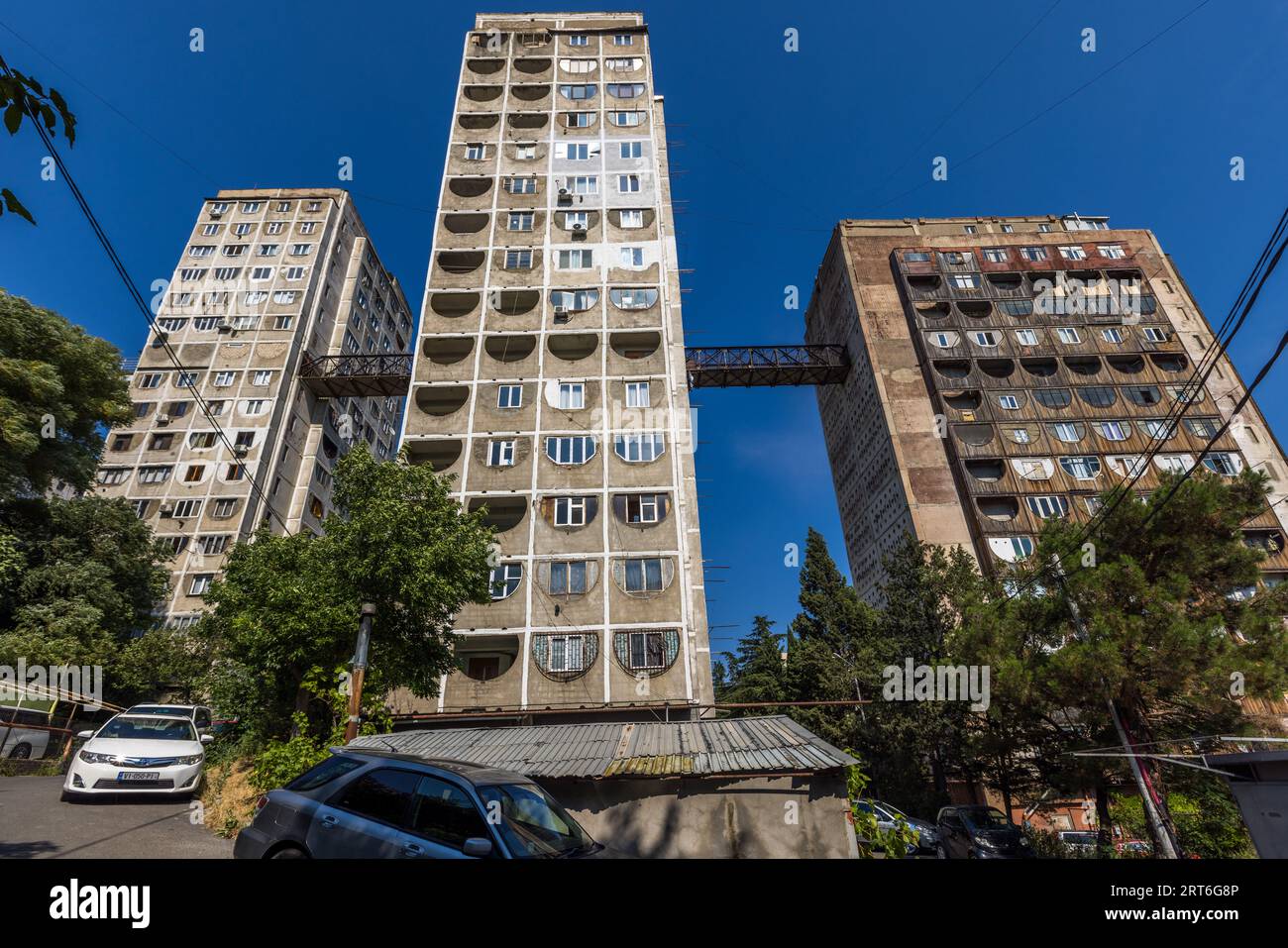 Il complesso residenziale Tbilisi Skybridge (Nutsubidze Skybridge) in via Ermile Bedia a Tbilisi, Georgia, si erge nel profondo cielo blu con tre blocchi di torri. Due passerelle a griglia arrugginite collegano la torre centrale ai suoi vicini a sinistra e a destra a un’altezza vertiginosa. La torre destra presenta elementi di facciata a forma di ferro di cavallo in mogano, quella centrale aperture a forma di ferro di cavallo di colore chiaro. Le auto sono parcheggiate davanti accanto a un capannone con tetto di lamiera ondulata, con alberi che crescono a sinistra e a destra. Il complesso collega i quartieri di Saburtalo e Nutsubidze sulla collina. Si trova a circa 11 kil Foto Stock