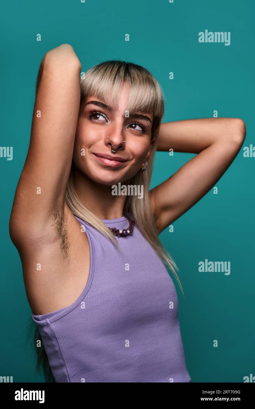 Giovane donna con capelli biondi e piercing che mette le braccia dietro la testa, mostrando l'ascella pelosa su sfondo turchese concetto positivo del corpo Foto Stock