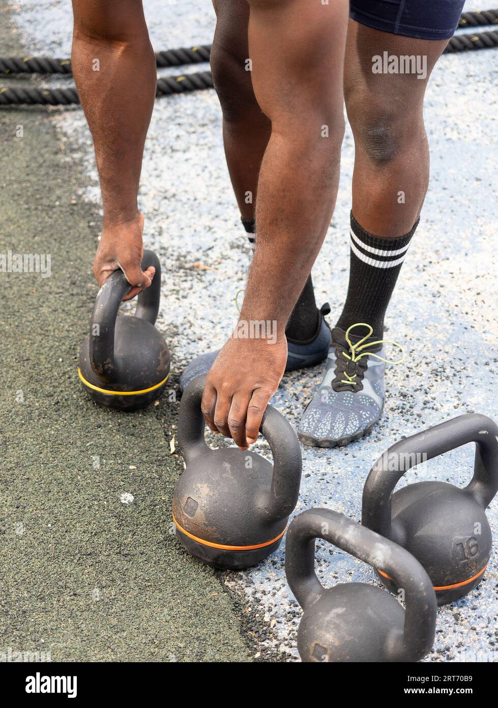 Dall'alto, anonimo uomo etnico in sneakers, mentre si piega in avanti e si schiaccia kettlebell a terra con le mani durante l'allenamento di giorno Foto Stock