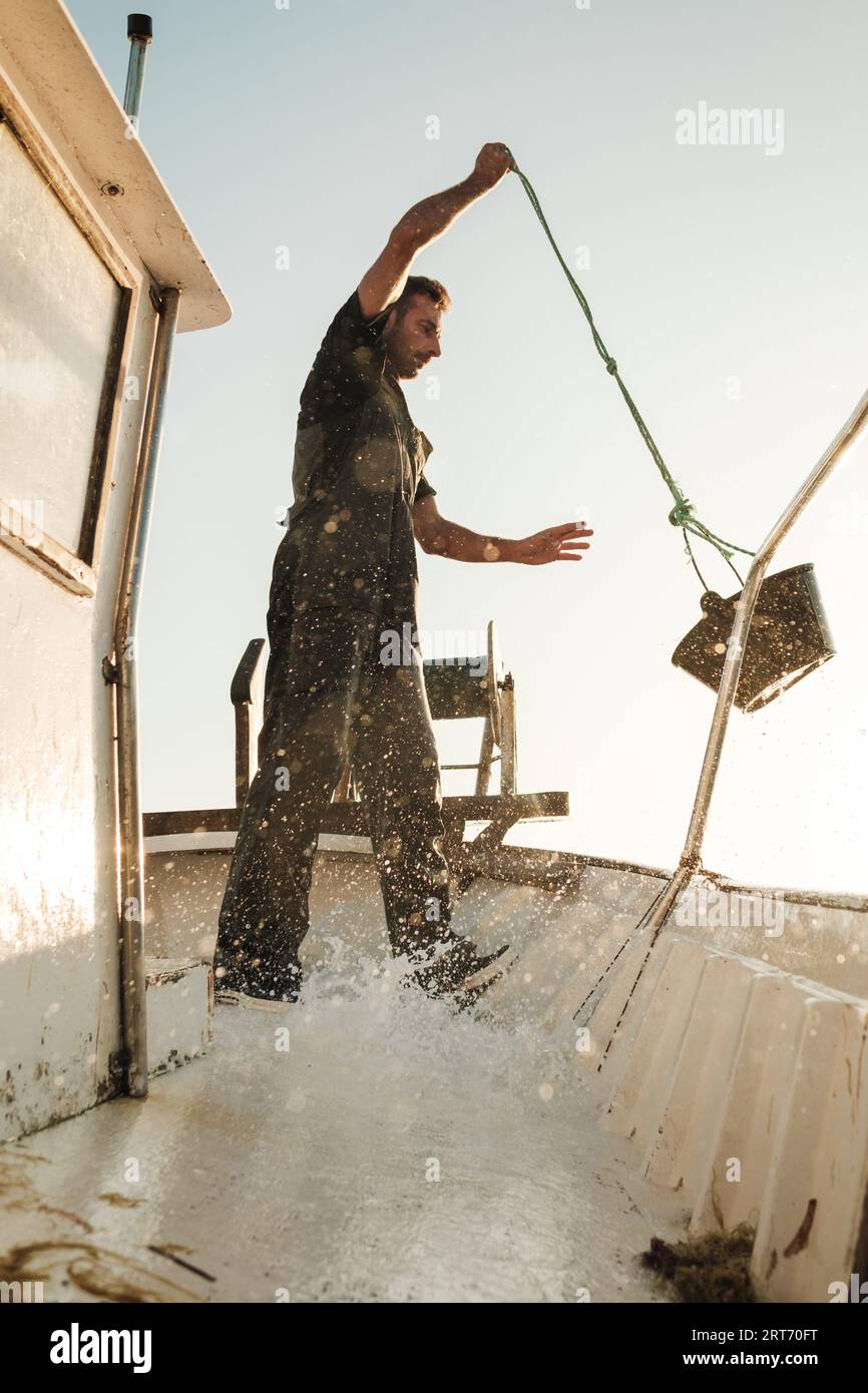 Angolo basso di pescatore maschio barbuto che versa acqua dal secchio sul ponte sporco della goletta mentre pesca a Soller, vicino all'isola delle Baleari di Maiorca Foto Stock