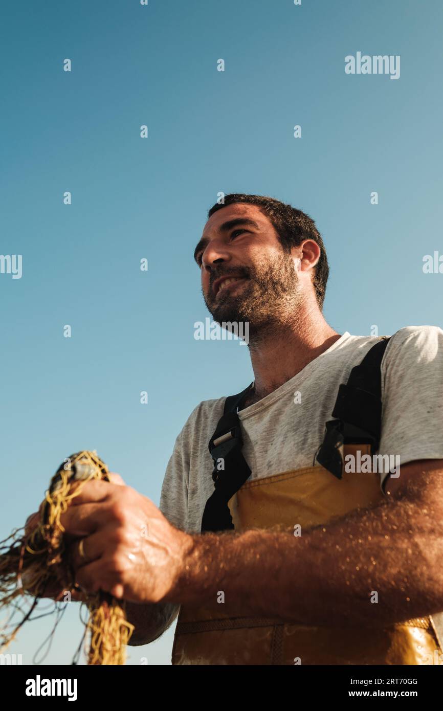 Sotto il ritratto di un felice marinaio maschio in grembiule con gli scontri da pesca ammirando il paesaggio marino mozzafiato al tramonto su una barca a vela a Soller, sull'isola delle Baleari Foto Stock