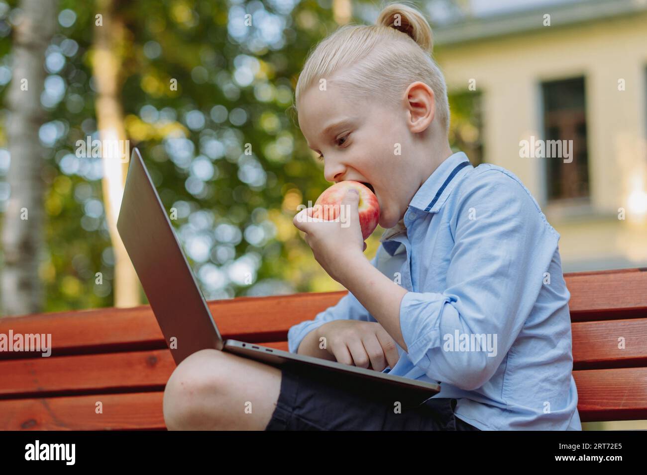 grazioso ragazzo caucasico con coda di cavallo seduto sulla panchina nel parco con un computer portatile che mangia mela. Fare i compiti all'aperto. Immagine con messa a fuoco selettiva Foto Stock