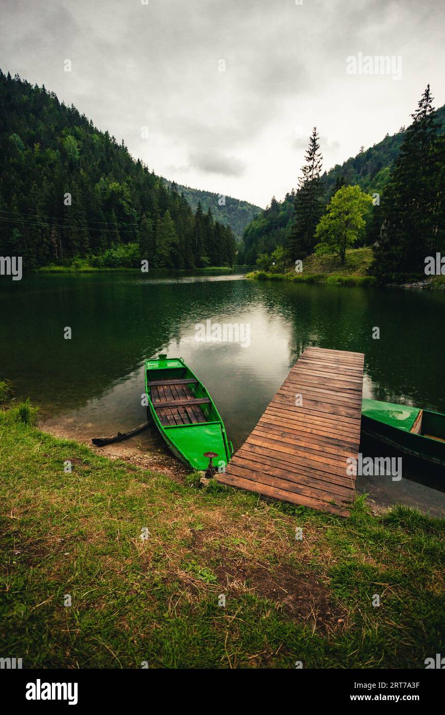 Foto di paesaggio verticale del bellissimo lago con talpa di legno e barche verdi al tramonto. Cielo drammatico e nuvoloso sul fiume che scorre attraverso la prua Foto Stock