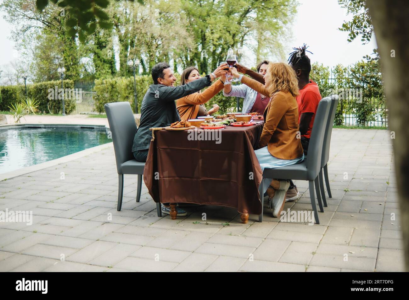 Un gruppo eterogeneo di amici, tra cui caucasici e un individuo africano, brindisi con bicchieri di vino in un cortile giardino vicino alla piscina. Si siedono togeth Foto Stock