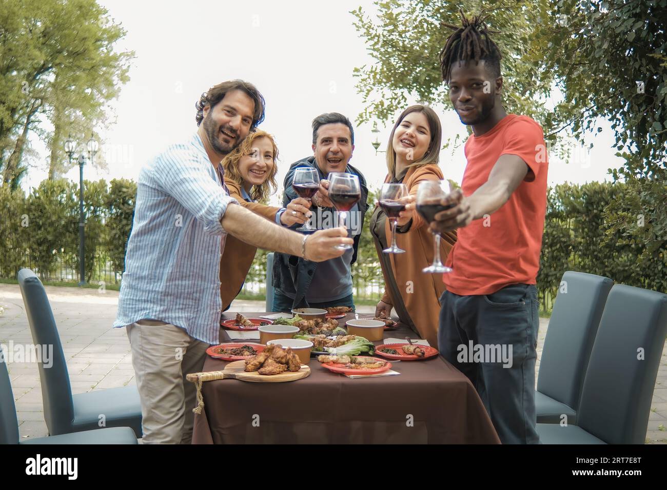 Un gruppo multietnico di amici si trova in un cortile giardino, alzando i bicchieri da vino per un brindisi. Guardando direttamente la telecamera, condividono sorrisi Foto Stock