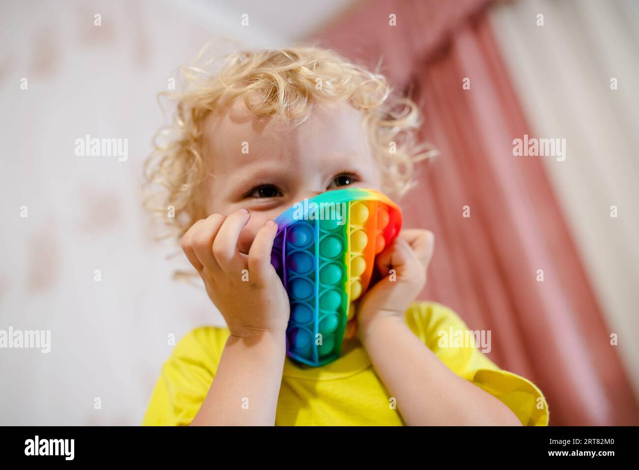 la ragazza si è ricoperta il viso con un rotondo multicolore. Bionda ragazza che gioca con pop it sensoriale giocattolo. gioco alla moda in silicone per bambini stressati Foto Stock