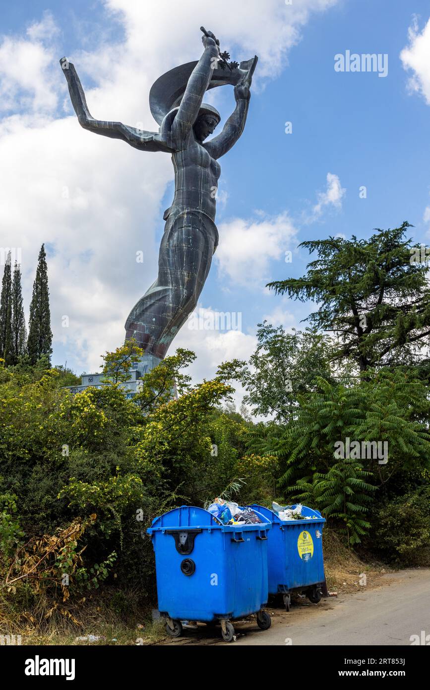 La statua di una donna si trova su un piedistallo presso il memoriale alla Tomba del Milite Ignoto a Vake, Tbilisi, Georgia, Europa. Le sue braccia sono alzate. Ha un ramo nella mano sinistra e una sciarpa nella mano destra. Il piedistallo è ricoperto di graffiti. Una scala conduce al piedistallo. Due bidoni blu pieni di rifiuti stanno in primo piano. Alberi e cespugli circondano la scena Foto Stock