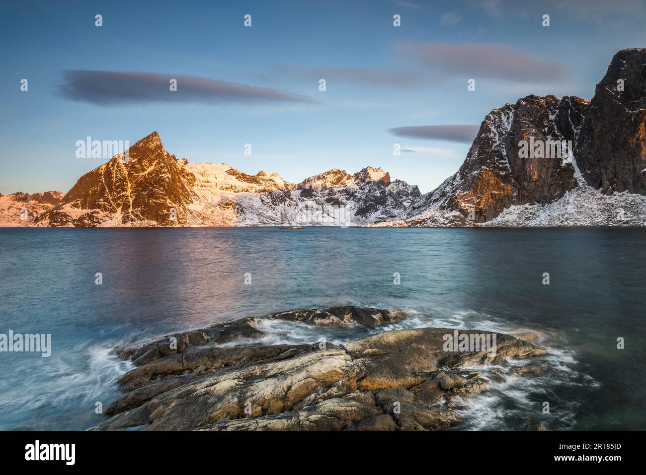 Calotte di ghiaccio su roccia scura con lago e ripide montagne innevate nel Reinefjord sulle isole Lofoten in Norvegia, nella luce dorata del mattino sul limpido Foto Stock