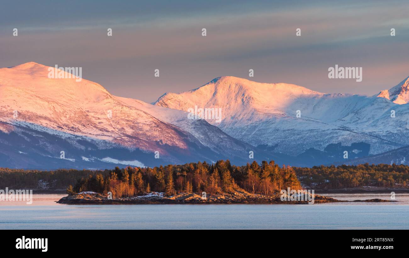 vista panoramica della costa norvegese con neve coperta montagne e piccola isola con alberi in bella serata morbida leggero Foto Stock