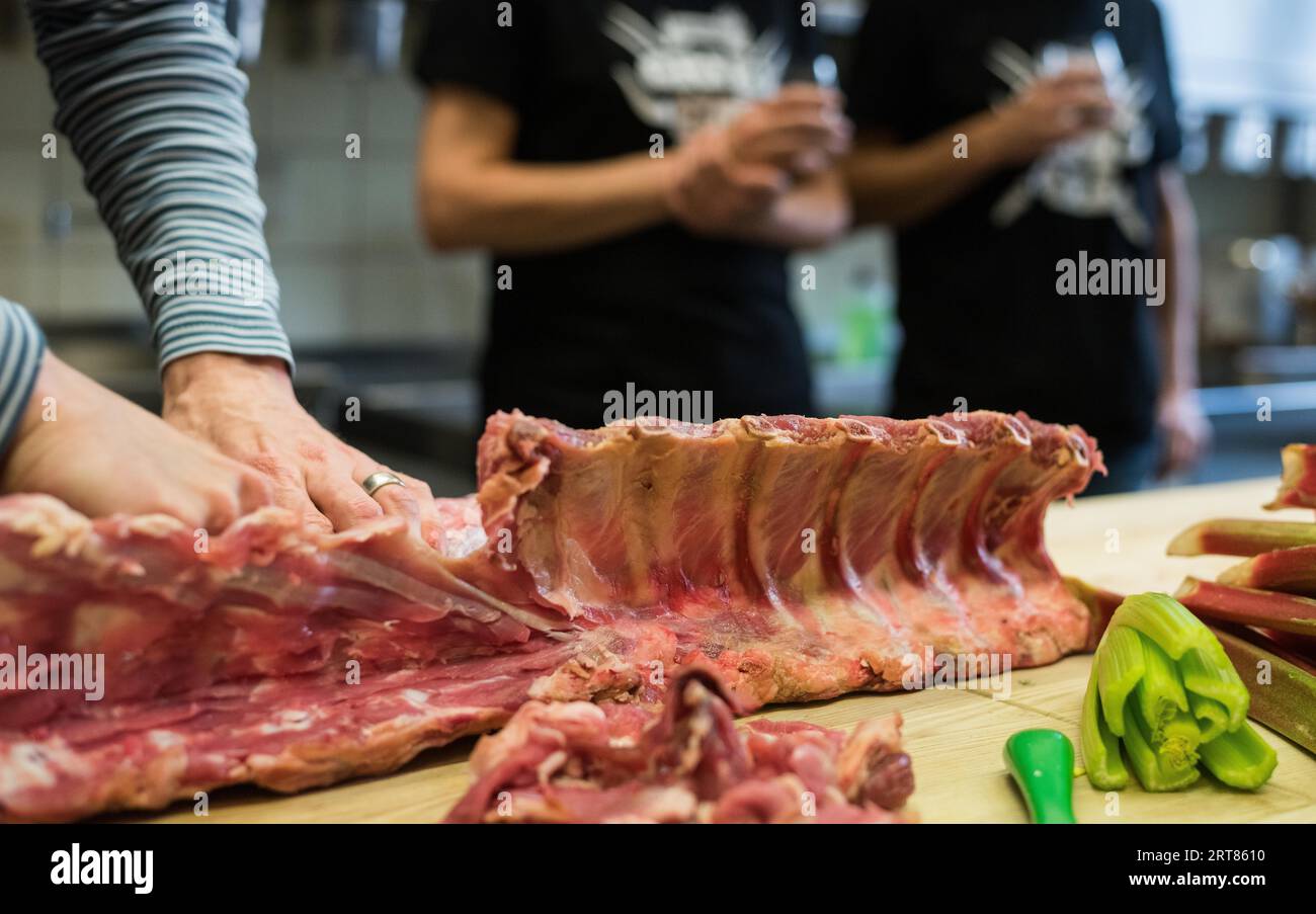 Mani umane di chef che tagliano rack di carne di agnello mentato su tagliere di cucina di legno con verdure e cucina in sfondo Foto Stock