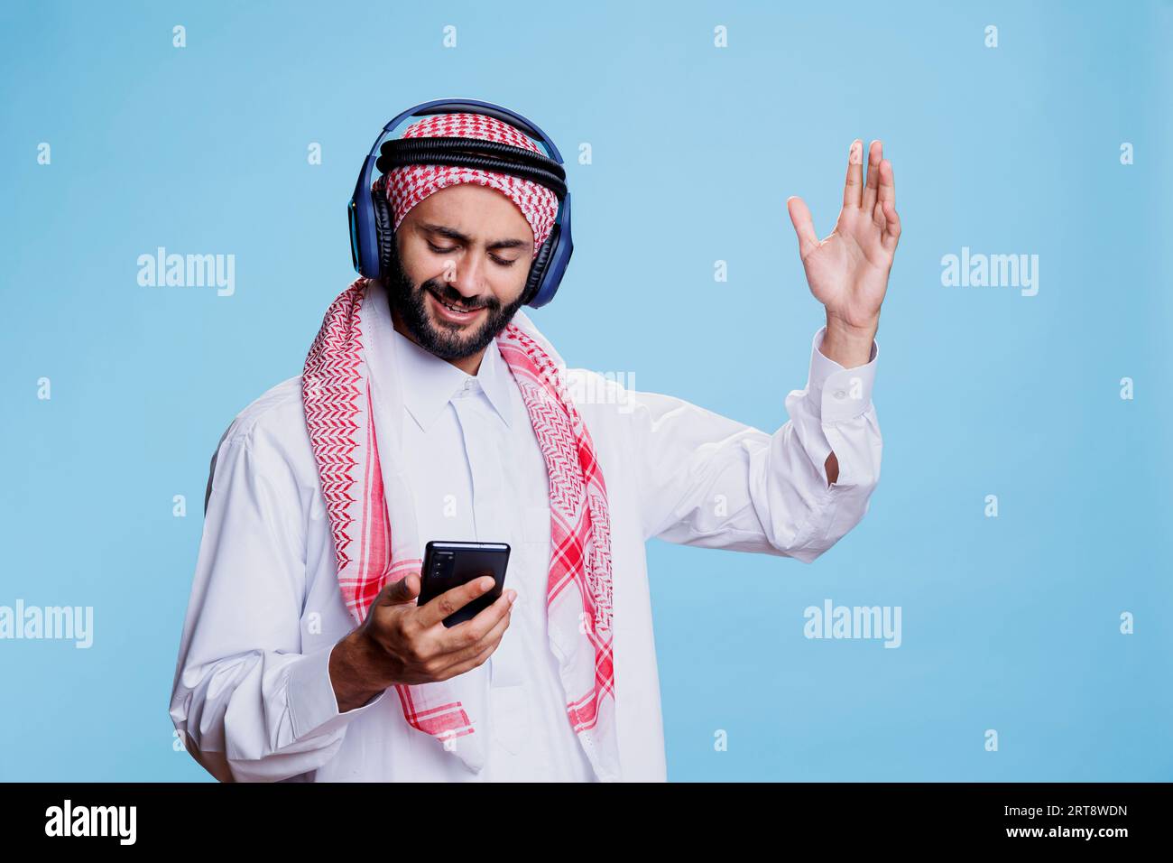 Uomo musulmano che indossa un velo ghutra e cuffie wireless che cambia canzone nella playlist sullo smartphone. Arabo ascolto di musica in cuffia, gesticolazione e utilizzo del telefono cellulare Foto Stock