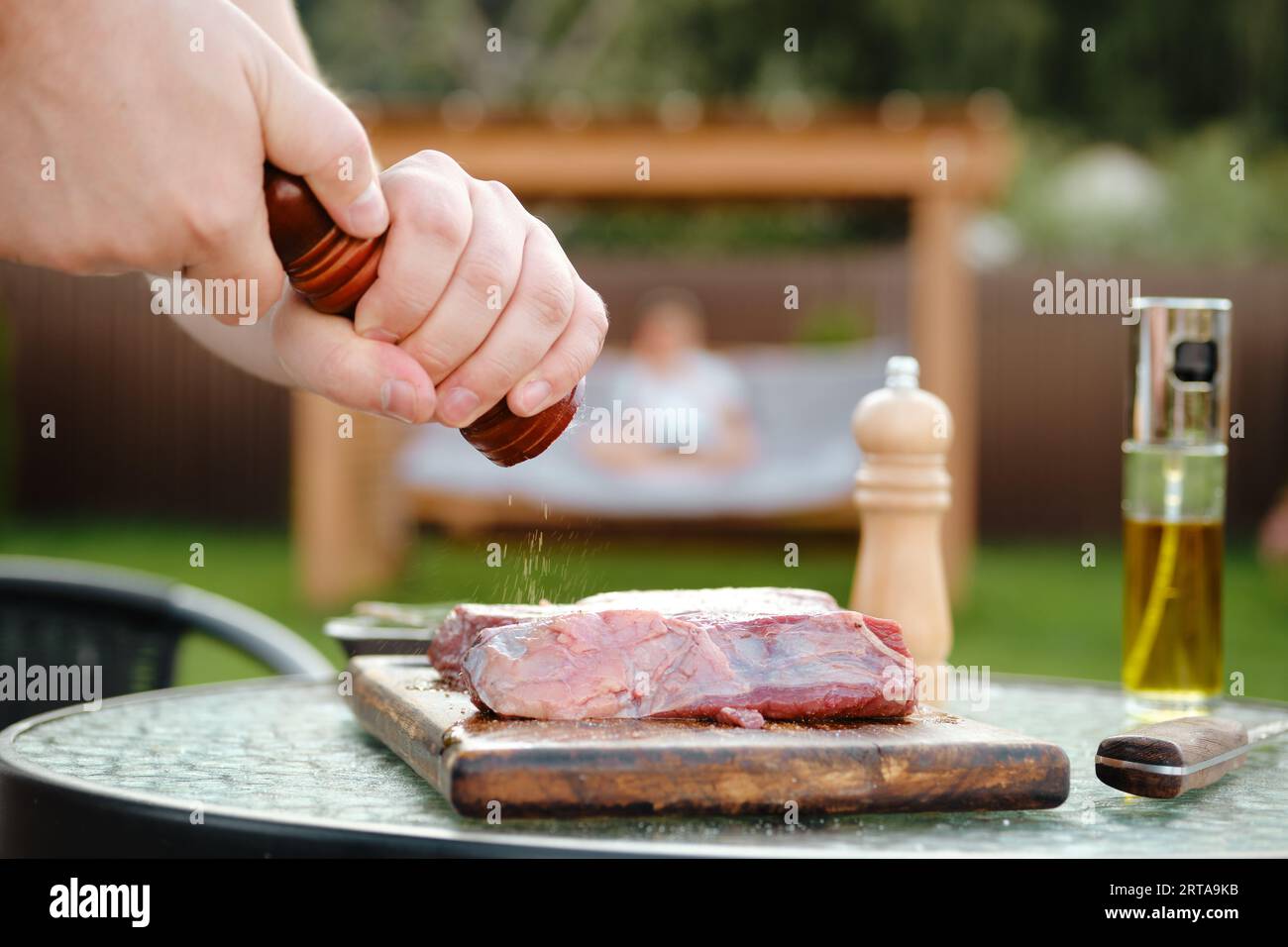 Vista laterale di un uomo irriconoscibile che aggiunge pepe alla bistecca di manzo crudo, che prepara succose carni alla griglia per cena con sua moglie Foto Stock