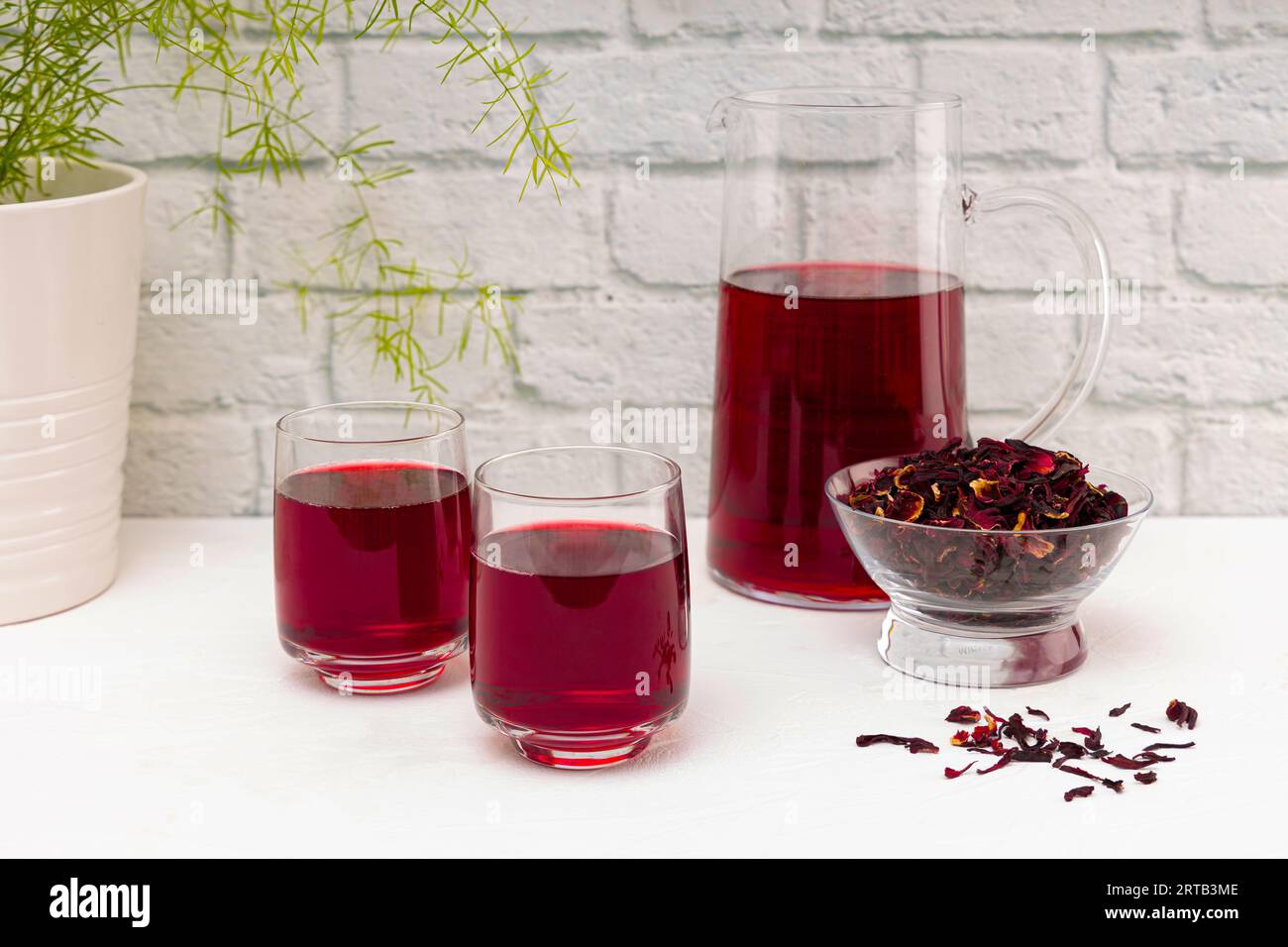 Brocca piena di tè secco con fiori di ibisco, su un tavolo di legno bianco. Foto Stock