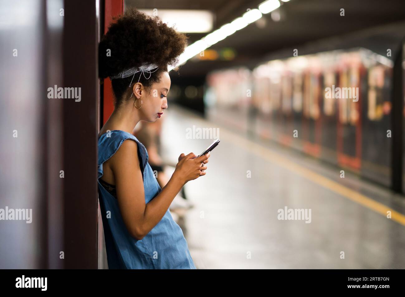 Vista laterale della giovane donna marocchina con fascia per capelli afro che guarda lo schermo del telefono cellulare mentre si è in piedi a controllare i messaggi di testo sulla piattaforma della metropolitana Foto Stock