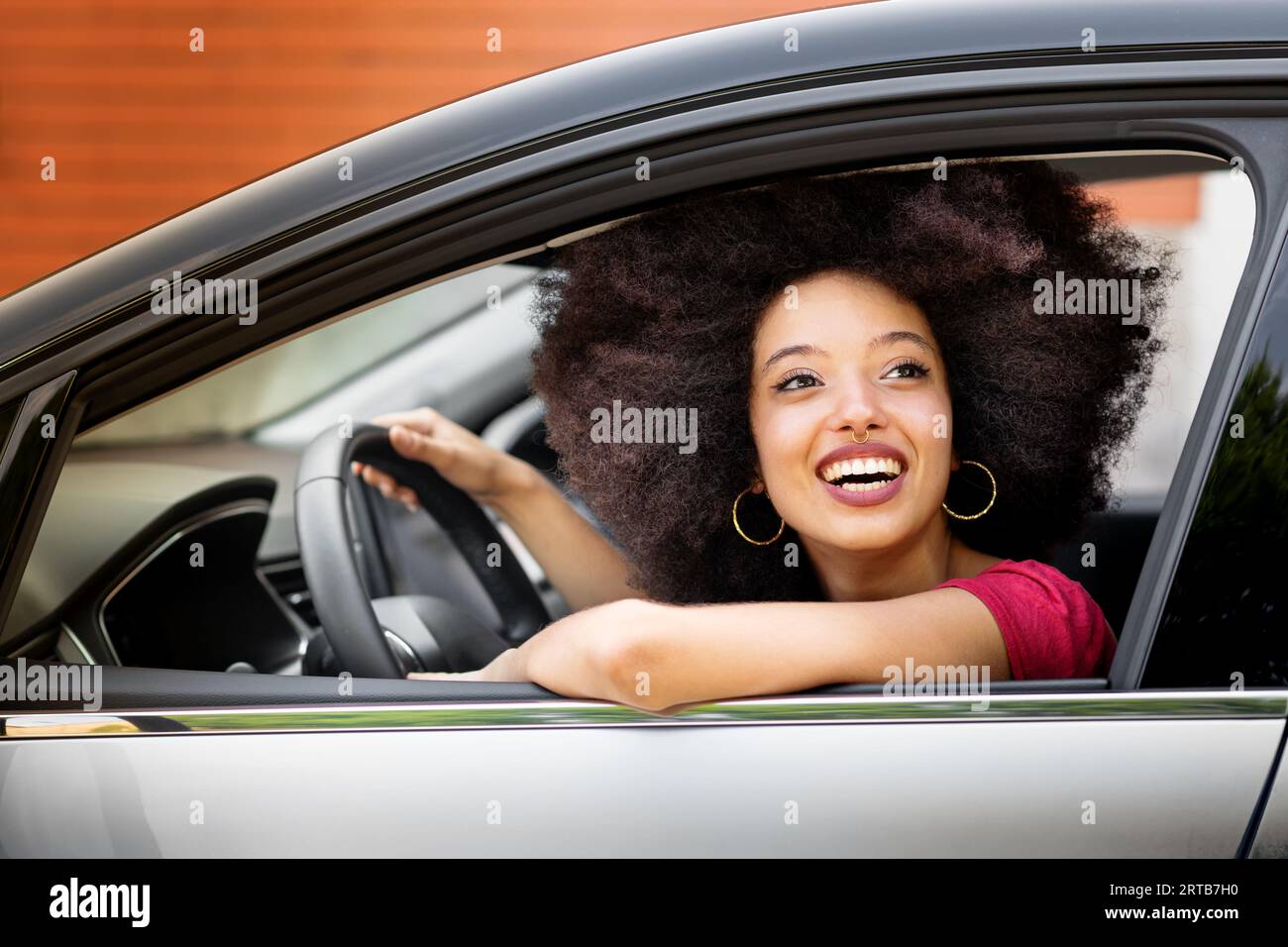 Giovane donna marocchina sorridente con acconciatura afro che guarda lontano mentre si siede sul sedile del conducente di un'auto parcheggiata di giorno con la mano sul volante e. Foto Stock